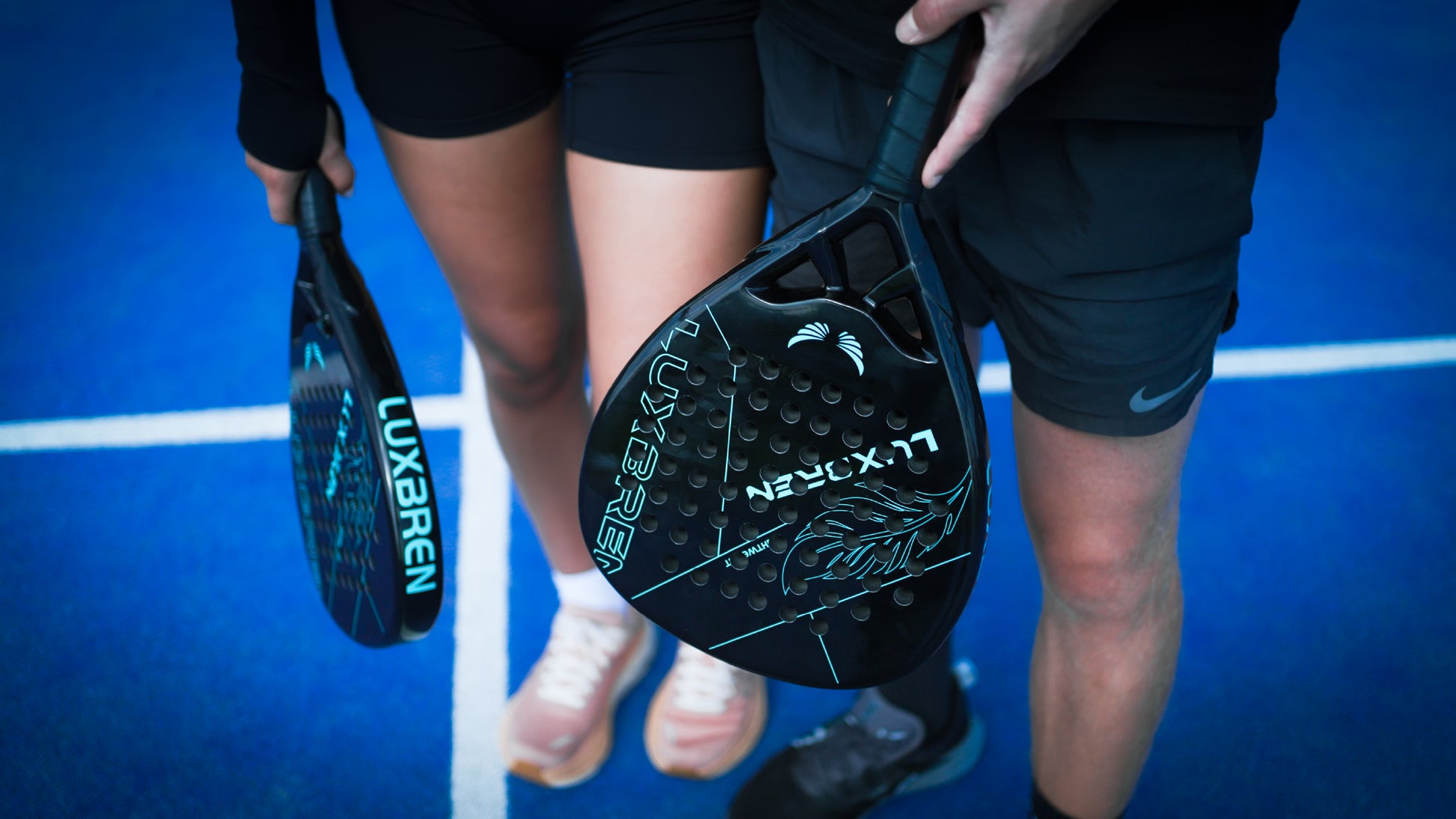 Man and woman holding Luxbren padel rackets on a blue outdoor padel court.