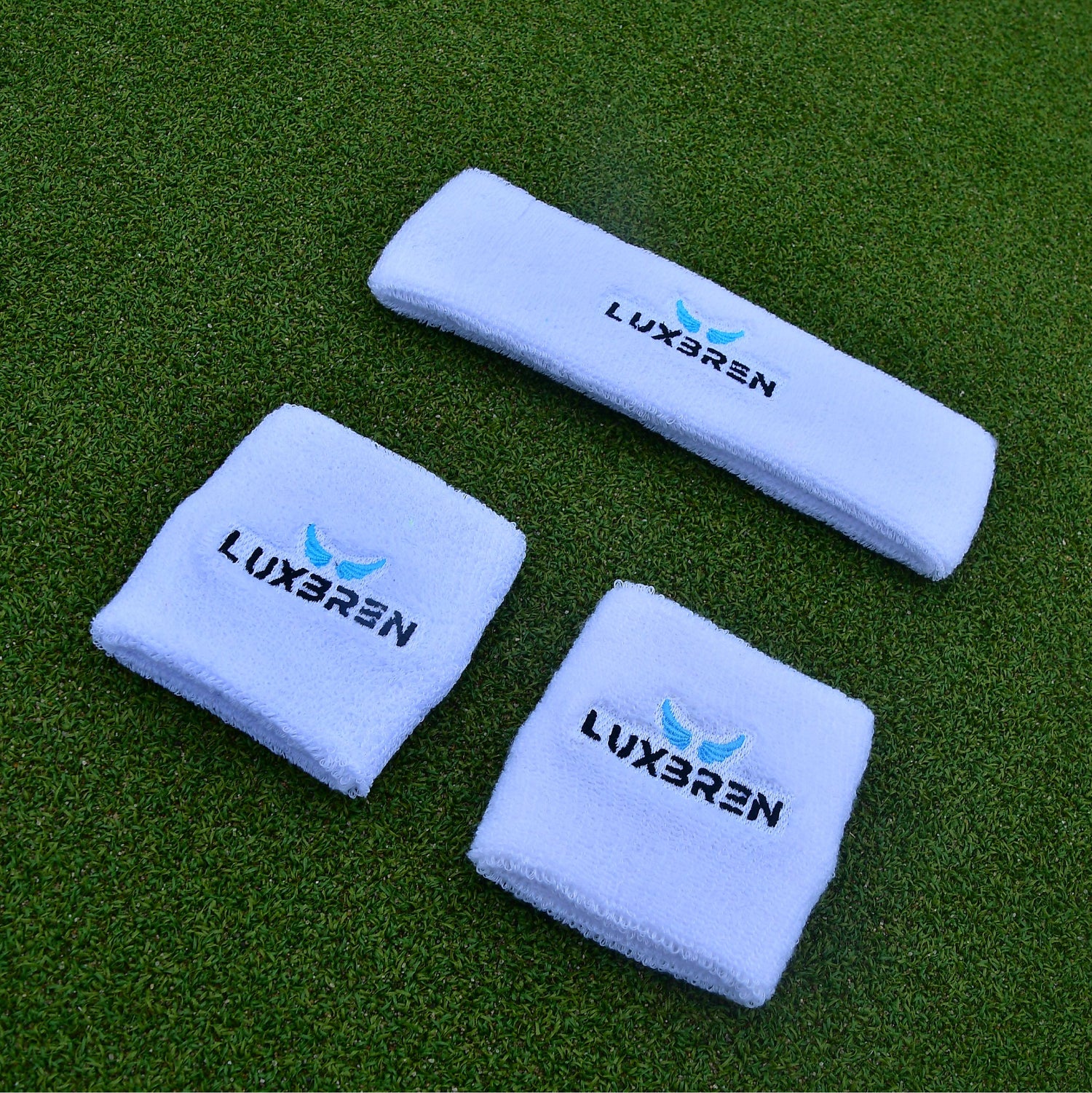 2 white sweatbands and 1 white headband with Luxbren blue and black branding on an outdoor tennis court